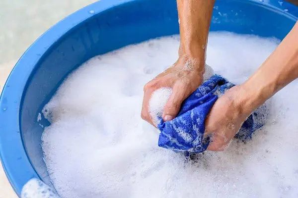 Đôi tay đang giặt một chiếc áo xanh trong chậu nước có bọt xà phòng.